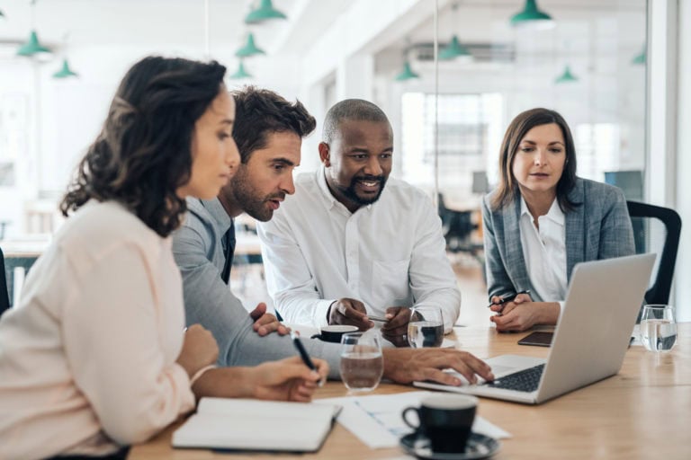 Diverse group of smiling businesspeople working together over a laptop during a meeting in the boardroom of a modern office