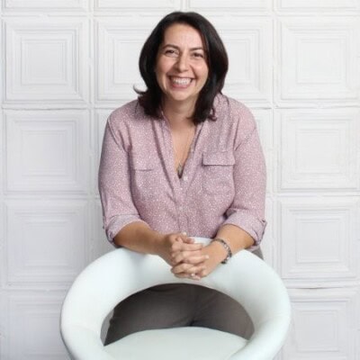 A person with dark hair, wearing a patterned blouse, is seated on a white chair, smiling, against a white paneled background.