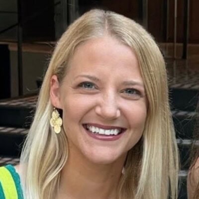 A smiling person with long blonde hair is wearing a green top and gold earrings, standing indoors.