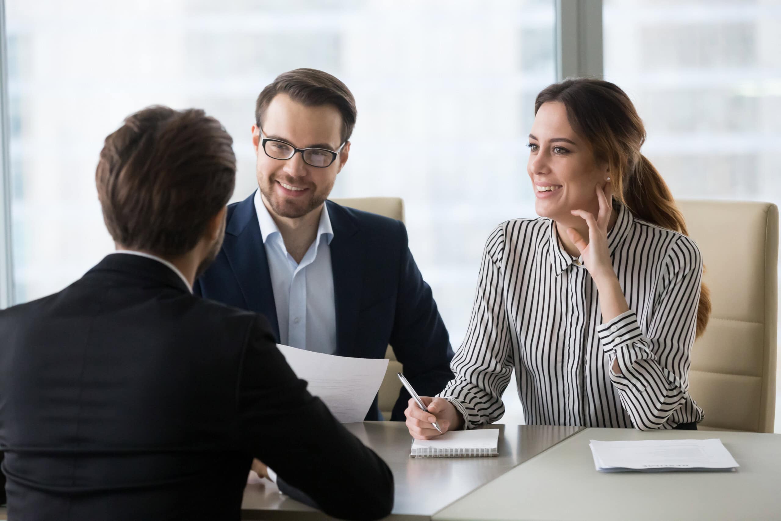 Three people are seated around a table in a modern office setting, engaged in a discussion. Two of them are facing the third person, smiling and holding papers.