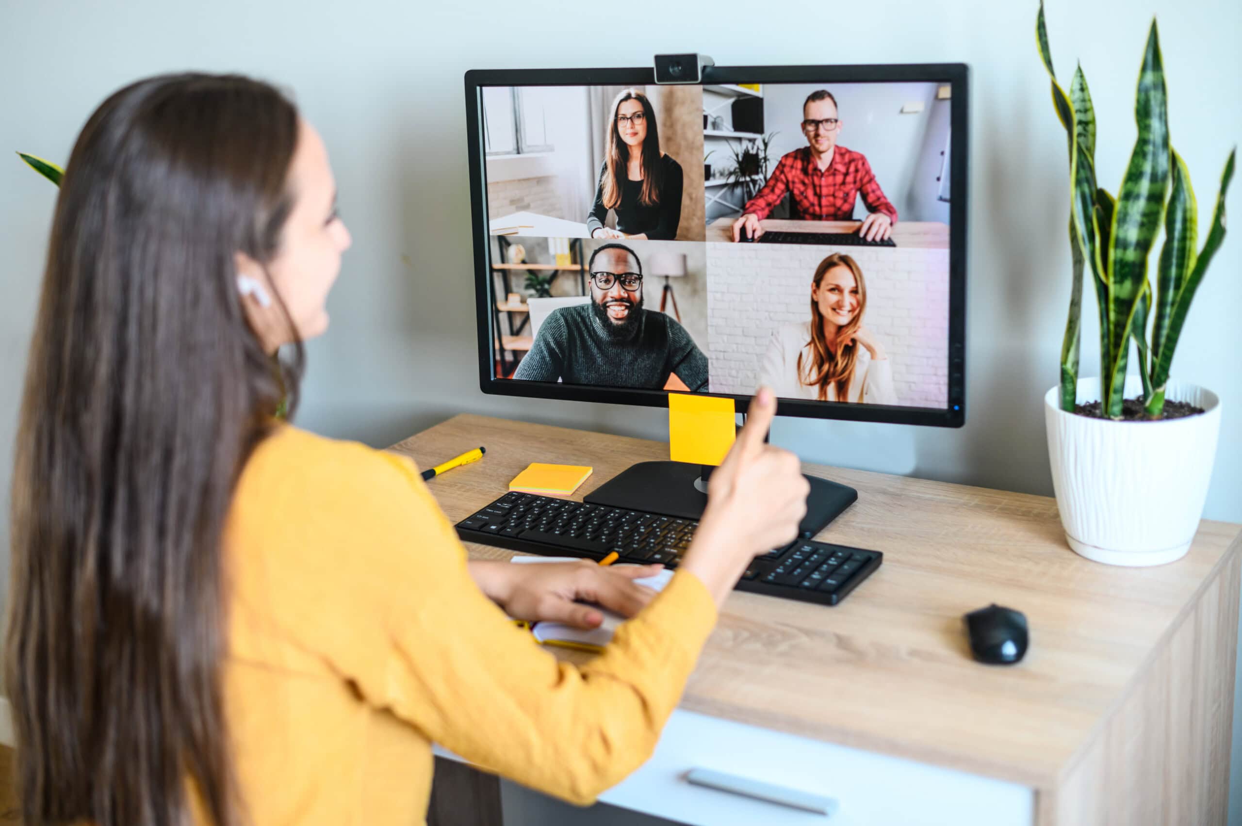 A woman engages in a video call, conducting online marketing research with four participants displayed on her computer monitor, surrounded by office supplies and a potted plant on her desk.