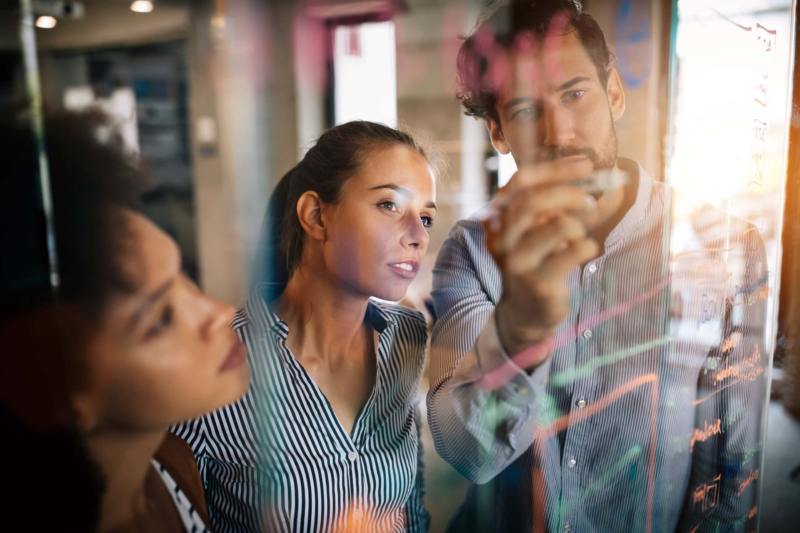 Three people collaborate, writing on a transparent board covered with colorful diagrams in a modern office setting.