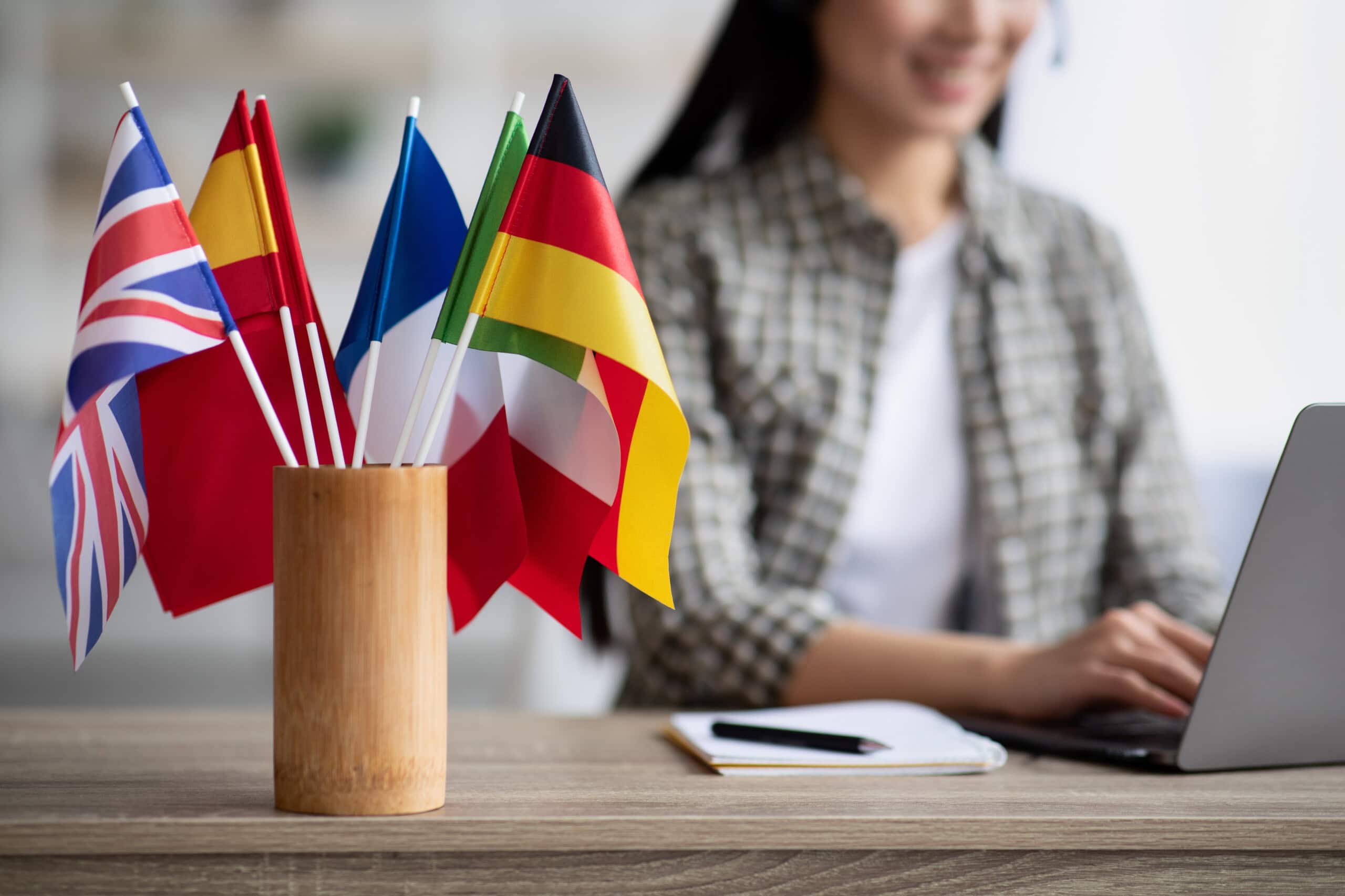 Small international flags in a wooden holder on a desk with a person typing on a laptop in the background.