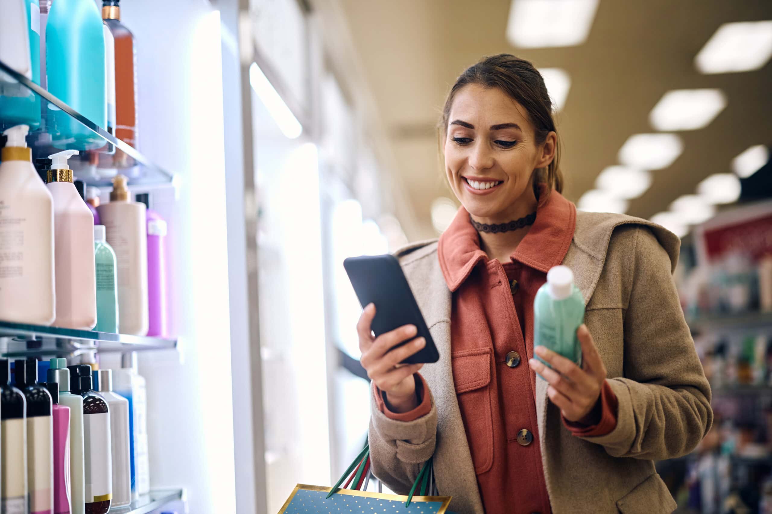 Woman checks her phone while holding a shampoo bottle in a store aisle, surrounded by various hair products.