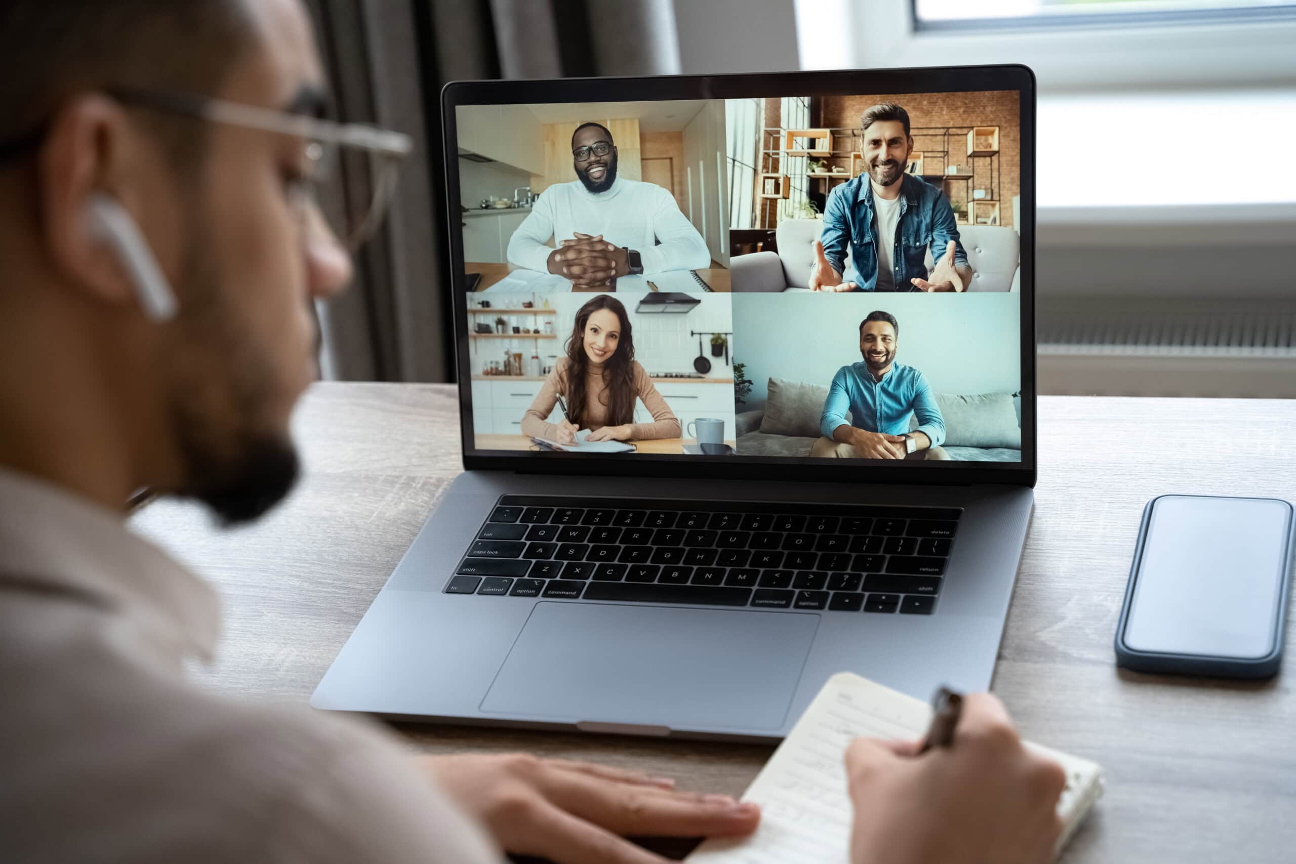 A person takes notes while participating in a video conference with four people displayed on a laptop screen.