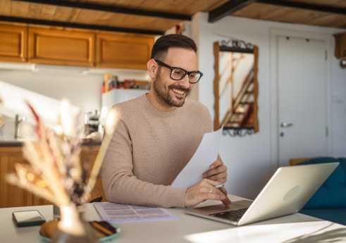 A man with glasses sits at a table in a cozy home setting, reviewing papers and smiling at his laptop, accessing a market research platform. Wooden cabinets and a staircase are visible in the background.