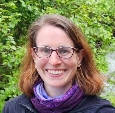 A woman with glasses and a purple scarf smiles outdoors with green foliage in the background.