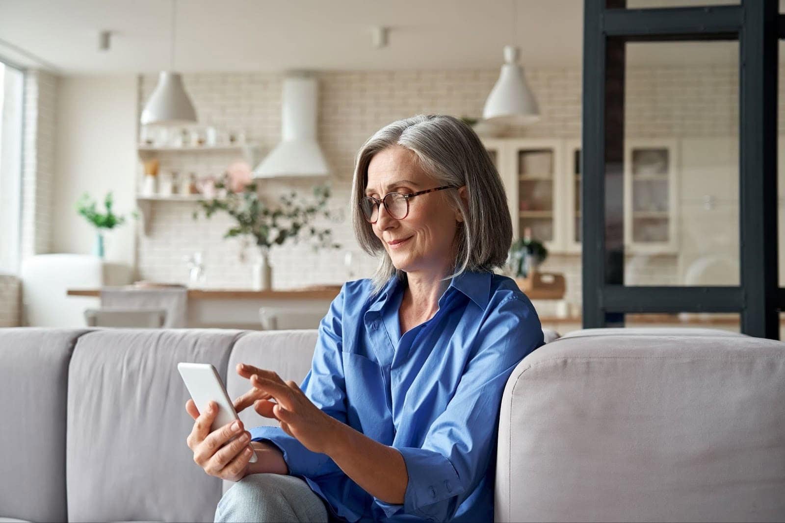 A woman with glasses and gray hair sits on a sofa using a smartphone in a modern living room.