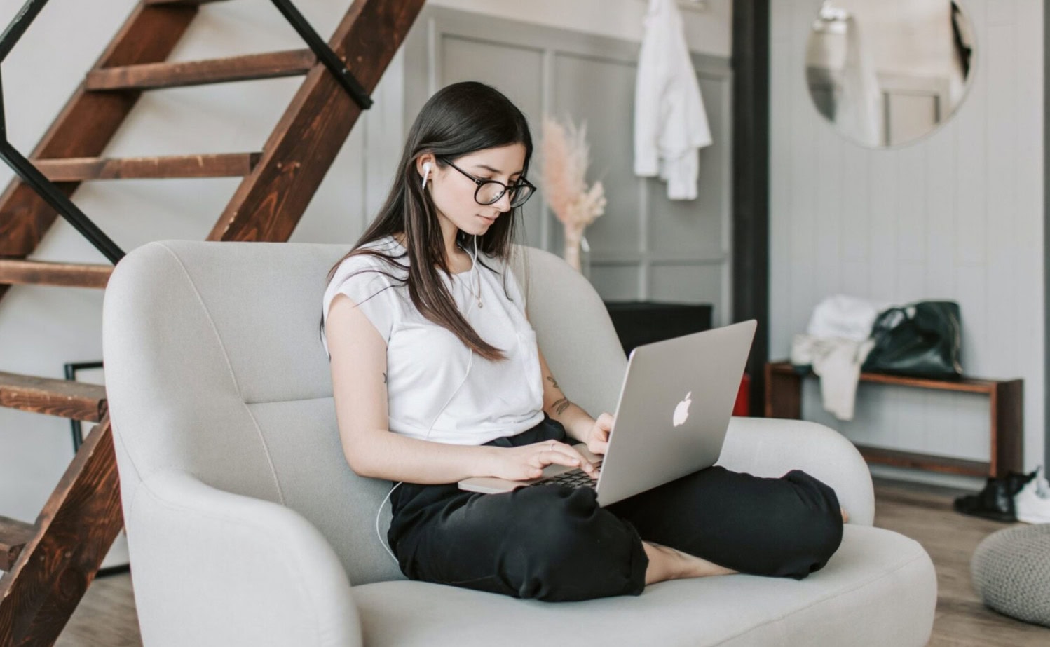 Person sitting on a couch, using a laptop, wearing earphones, in a modern living room with a staircase in the background.