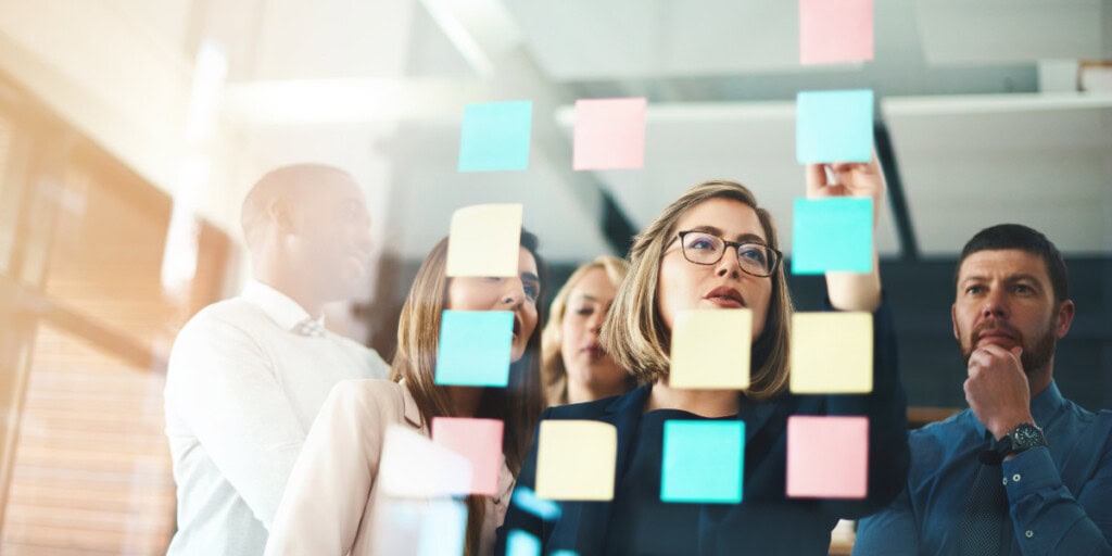 A group of people in an office setting are placing sticky notes on a glass wall while discussing.