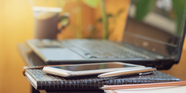 A smartphone, pen, and notebook next to a laptop and coffee cup on a small table.