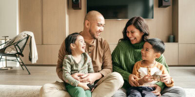 A family of four sits on the floor in a living room, smiling and laughing together. The parents each hold one of their children.